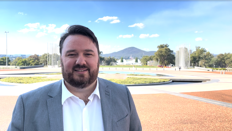 The image features Chris Armstrong, who is in an outdoor setting with a mountainous landscape in the background. He is wearing a suit and standing on a paved area surrounded by greenery and modern structures.