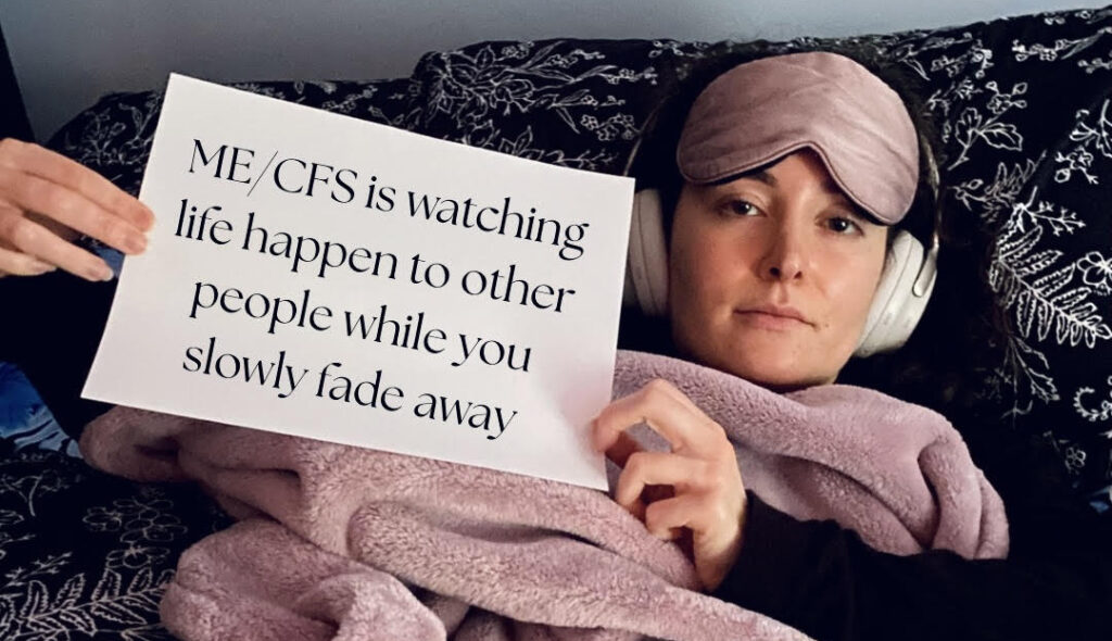 Samantha is holding a sign that reads: ME/CFS is watching life happen to other people while you slowly fade away.